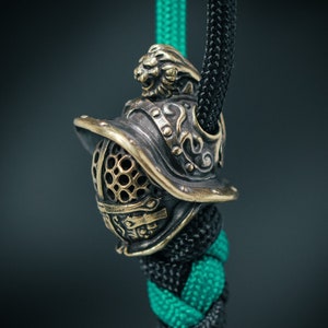 May include: A bronze gladiator helmet bead with a lion head detail, hanging on a green and black paracord.