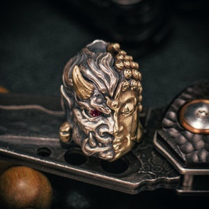 May include: A silver and gold metal charm featuring a detailed depiction of a demon and a Buddha face. The demon has a red gemstone eye and the Buddha has a serene expression.
