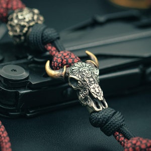 May include: A red and black paracord bracelet with a silver and gold bull skull charm. The charm is detailed with floral patterns and a face.