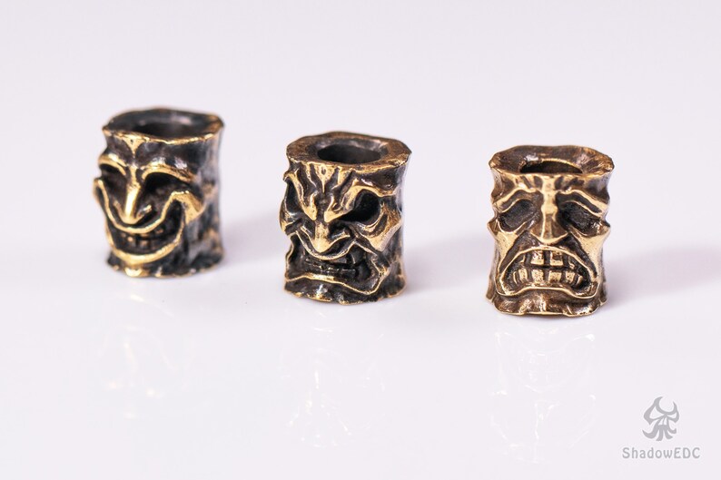 May include: Three brass beads with different facial expressions. Each bead features a detailed, textured design of a face with a unique expression. The beads are a dark brass color with a gold finish.