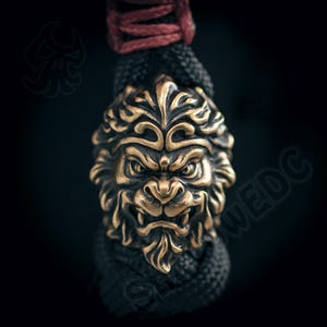 Pode incluir: Um pendente bronzeado em forma de cabeça de leão, com uma juba detalhada e uma expressão feroz. Está pendurado num cordão preto entrançado, com uma secção vermelha no topo. O fundo escuro realça os detalhes do pendente.