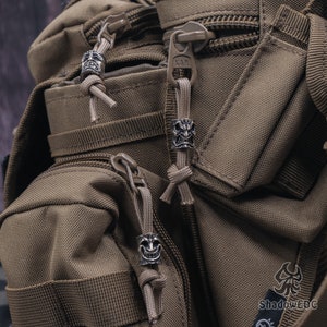 May include: Close-up of a brown tactical backpack with three silver skull-shaped zipper pulls. The backpack has a zippered pocket on the front and a large main compartment.