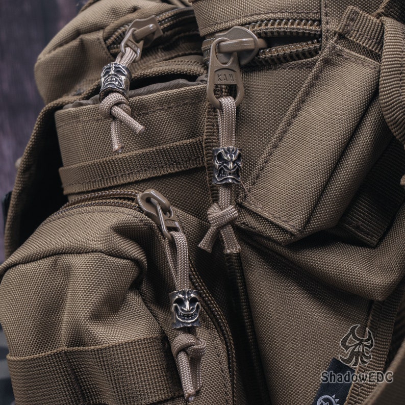 May include: Close-up of a brown tactical backpack with three silver skull-shaped zipper pulls. The backpack has a zippered pocket on the front and a large main compartment.