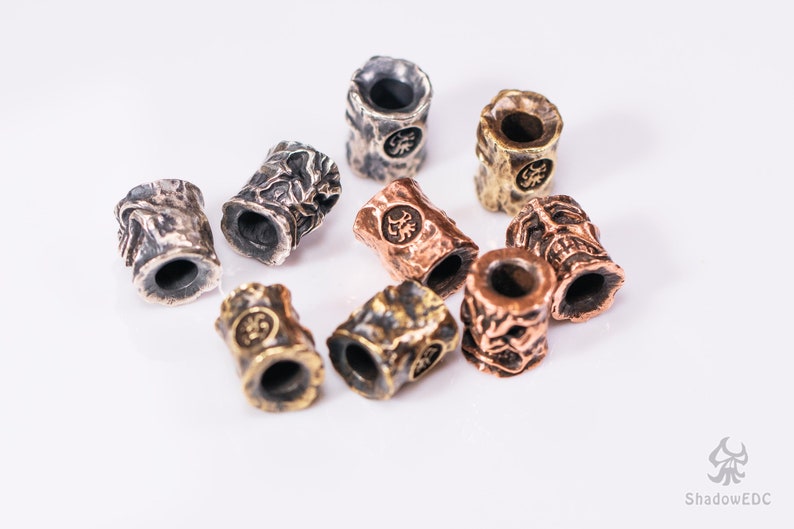May include: A collection of eight metal beads with intricate designs. The beads are in various shades of silver, gold, and copper. Each bead has a unique pattern and a small hole in the center.