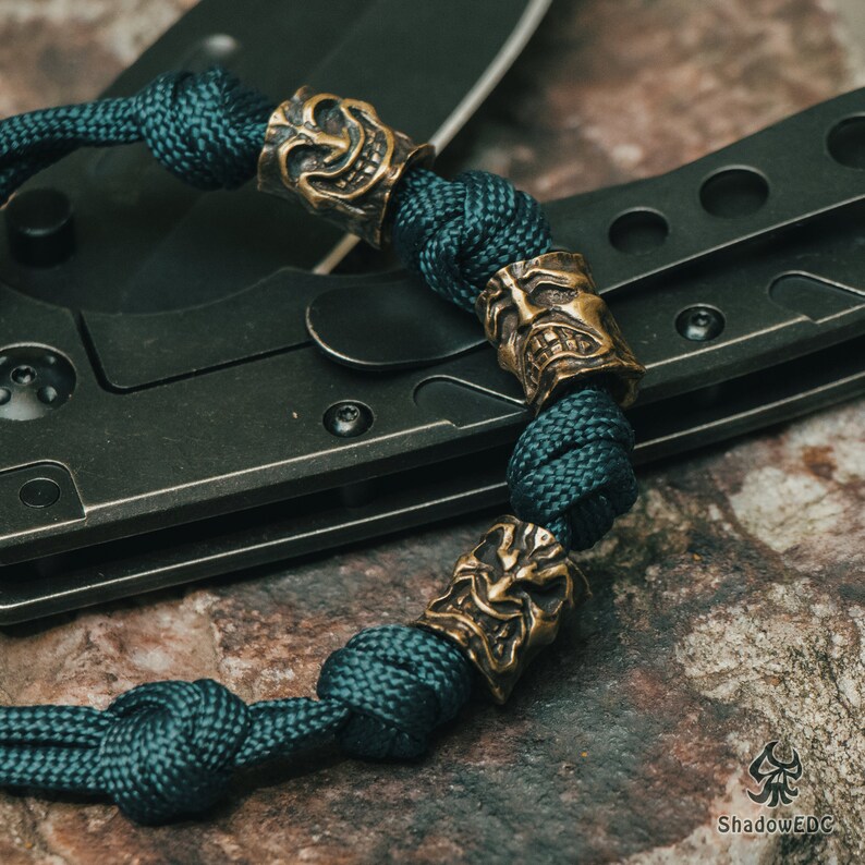 May include: A dark green paracord bracelet with bronze skull beads. The bracelet is wrapped around a black folding knife.