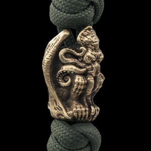 Könnte beinhalten: Eine bronzefarbene Perle mit einem detaillierten Design einer mythischen Kreatur. Die Perle hängt an einem dunkelgrünen Seil, mit Tentakeln und einem detaillierten Gesicht. Der Hintergrund ist schwarz.