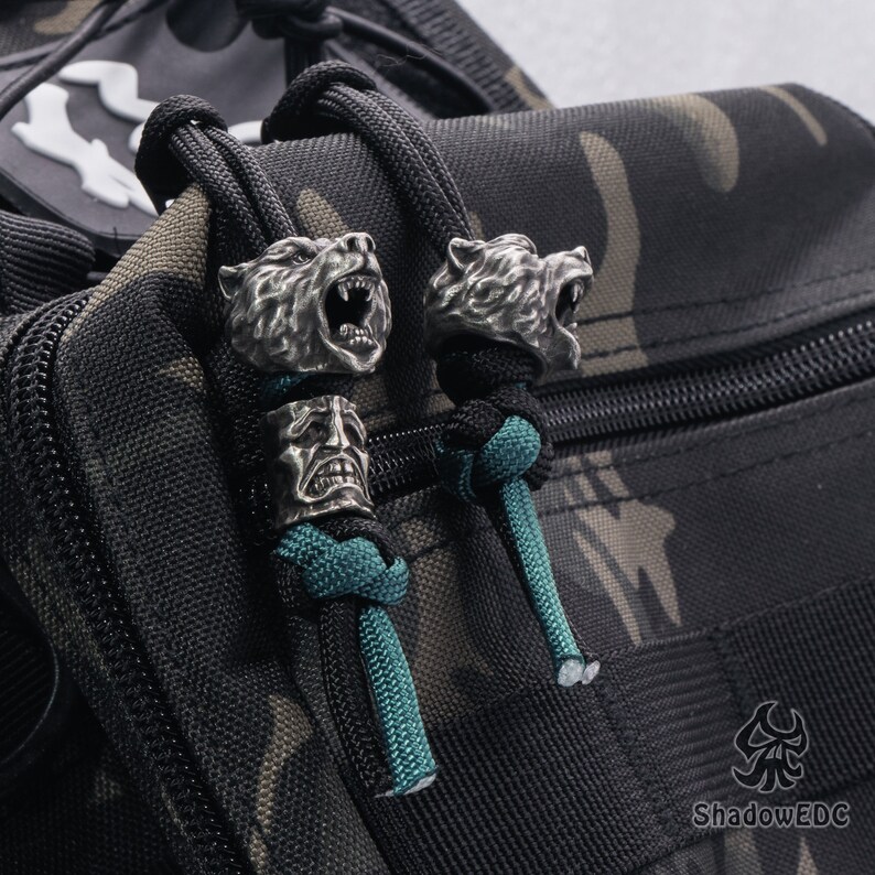 May include: Two silver bear head zipper pulls with teal paracord attached to a black and tan camouflage bag. The bag has a zipper and a black strap with the text "ShadowEDC" on it.