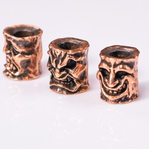 May include: Three copper-colored metal beads with a detailed, textured design of a face with a wide, toothy grin. The beads are arranged in a row on a white surface.