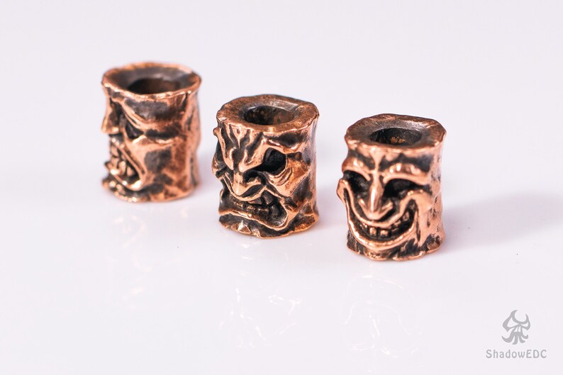 May include: Three copper-colored metal beads with a detailed, textured design of a face with a wide, toothy grin. The beads are arranged in a row on a white surface.