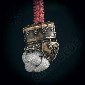 Peut inclure: Gros plan d'une perle en bronze en forme de casque de chevalier avec des détails complexes. La perle est attachée à un cordon rouge et blanc.