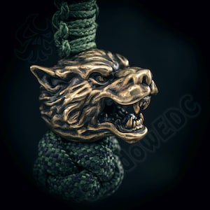 Puede incluir: Un colgante con cabeza de lobo de color bronce con detalles de pelaje y boca abierta, sujeto a un cordón trenzado verde y negro. El colgante es una toma de primer plano sobre un fondo oscuro, que muestra el intrincado diseño y la textura.