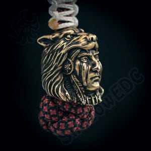 Puede incluir: Colgante de color bronce que representa a un jefe nativo americano con un tocado de lobo. El colgante está sujeto a un cordón trenzado blanco y rojo, con un nudo decorativo rojo y negro. El diseño es detallado e intrincado.