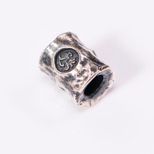 May include: A silver metal bead with a textured surface and a circular design featuring a scorpion. The bead has a hole through the center for threading.