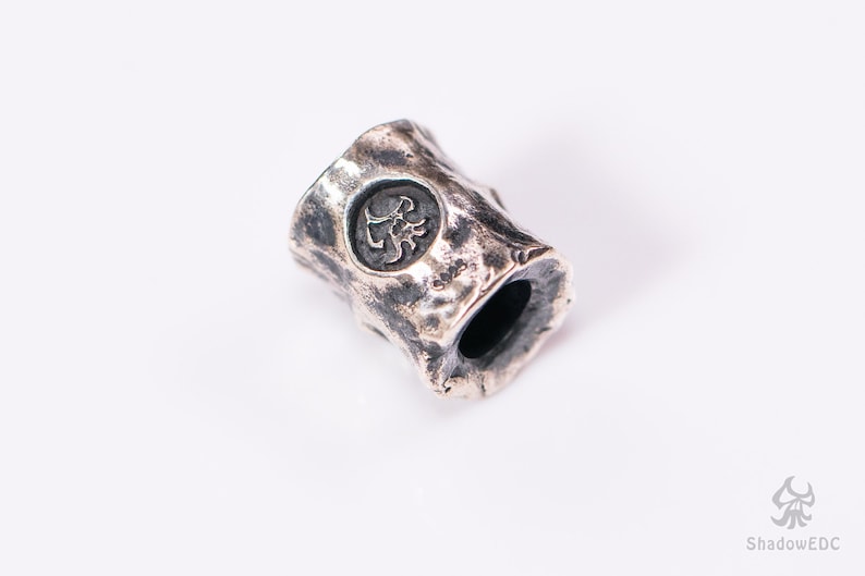 May include: A silver metal bead with a textured surface and a circular design featuring a scorpion. The bead has a hole through the center for threading.