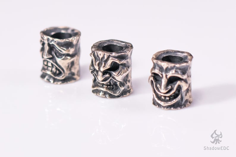 May include: Three silver-colored metal beads with detailed carvings of faces. Each bead features a different expression, including a frowning face, a grimacing face, and a smiling face.