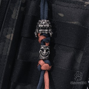 May include: A black and red paracord lanyard with two silver metal beads. The beads are shaped like a lion's head and a smiling face.