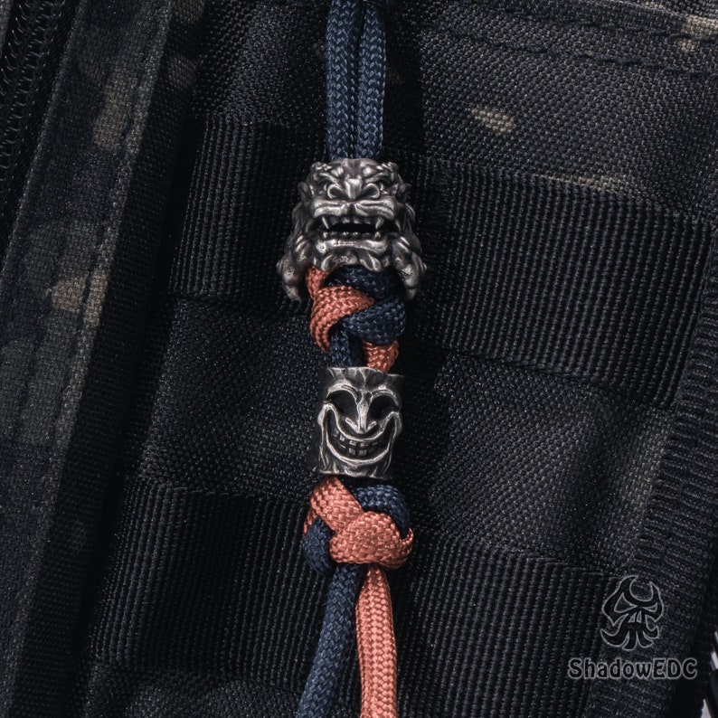 May include: A black and red paracord lanyard with two silver metal beads. The beads are shaped like a lion's head and a smiling face.