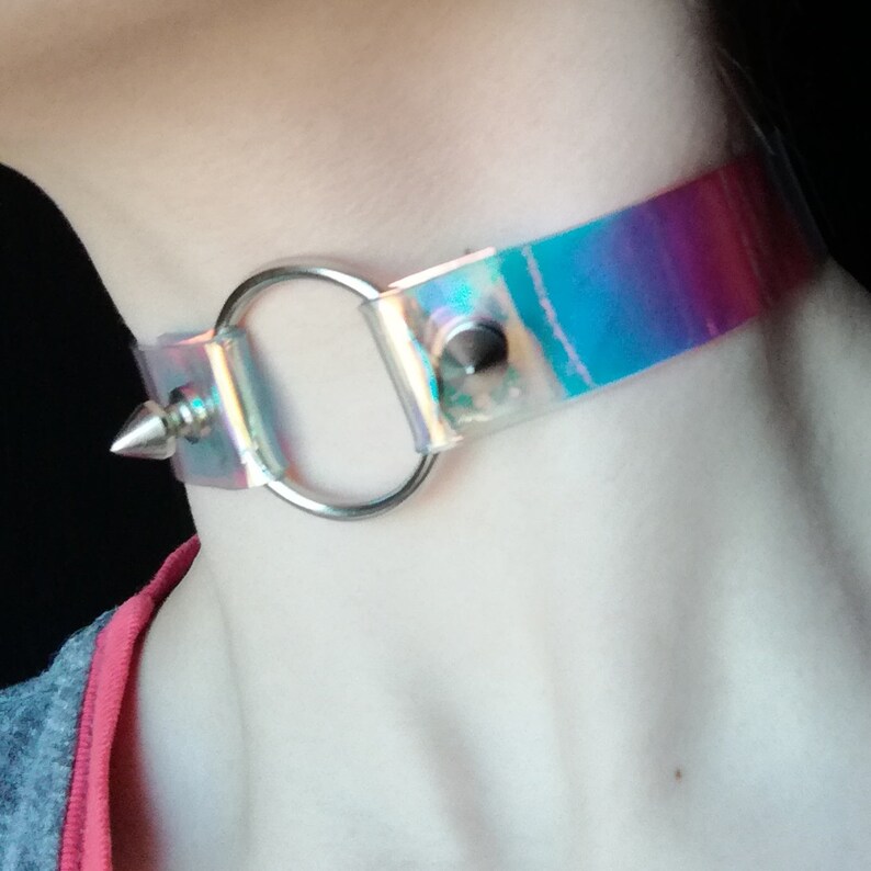 May include: A clear iridescent choker with a silver ring and a silver spike. The choker is made of a flexible material and has a rainbow iridescent finish.