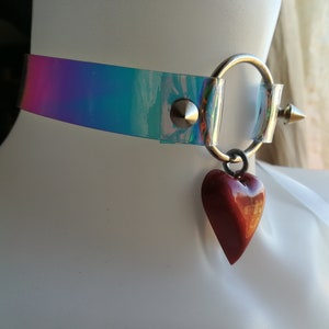 May include: A clear iridescent choker with a silver ring and a red heart charm. The choker has a silver spike on one side.
