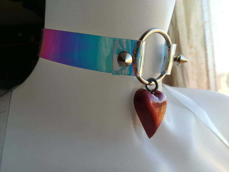 May include: A clear iridescent choker with a silver ring and a red heart charm. The choker has a silver spike on one side.