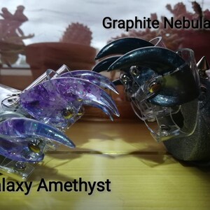 Puede incluir: Dos anillos en forma de garra con bases de pl&aacute;stico transparente. Un anillo est&aacute; decorado con purpurina morada y la etiqueta "Galaxy Amethyst". El otro anillo est&aacute; decorado con purpurina azul oscuro y la etiqueta "Graphite Nebula".