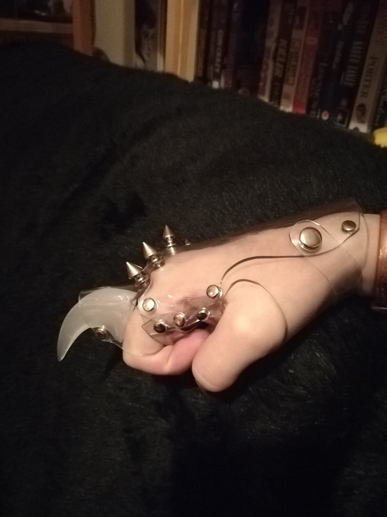 Puede incluir: Un anillo de garra de pl&aacute;stico transparente con p&uacute;as y tachuelas de metal plateado. El anillo se lleva en una mano.