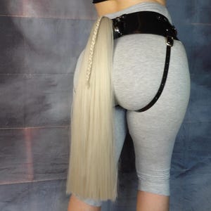 Può includere: Un'estensione di coda di capelli biondi intrecciati, attaccata a una cintura nera con un cinturino a bretella. La cintura è indossata sopra leggings capri grigi. L'estensione dei capelli è lunga e dritta.