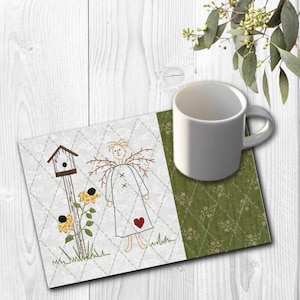 K&ouml;nnte beinhalten: Eine wei&szlig;e Keramiktasse steht auf einem gesteppten Stoffuntersetzer. Der Untersetzer zeigt ein gesticktes Design einer Vogelscheuchenfigur, eines Vogelhauses und Sonnenblumen. Der Untersetzer hat einen wei&szlig;en Hintergrund mit einem Rautenmuster und einem gr&uuml;nen Blumenmuster.