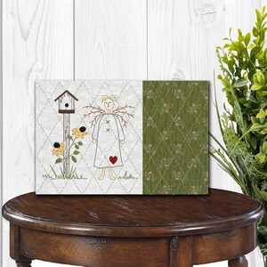 K&ouml;nnte beinhalten: Ein dekoratives rechteckiges Kunstwerk mit einem Folk-Art-Design. Die linke Seite zeigt einen gestickten Engel mit Herz, Vogelhaus und Sonnenblumen. Die rechte Seite hat ein gr&uuml;nes Blumenmuster. Das Kunstwerk wird auf einem dunklen Holztisch pr&auml;sentiert.