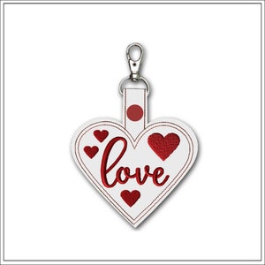 以下が含まれることがあります： 赤い刺繍の「love」と小さなハートがついた白いハート型のキーホルダー。キーホルダーにはシルバーのクリップが付いています。