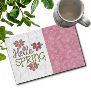 May include: A greeting card with the text "Hello SPRING" embroidered in green and gray, with pink flower accents. The card is divided into two sections, one white with a floral pattern and the other pink. A white mug is in the background.