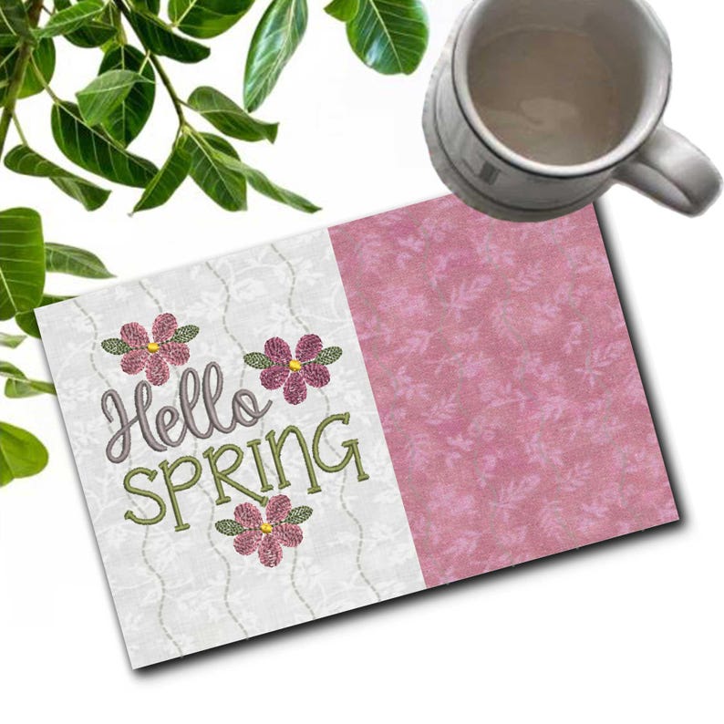 May include: A greeting card with the text "Hello SPRING" embroidered in green and gray, with pink flower accents. The card is divided into two sections, one white with a floral pattern and the other pink. A white mug is in the background.