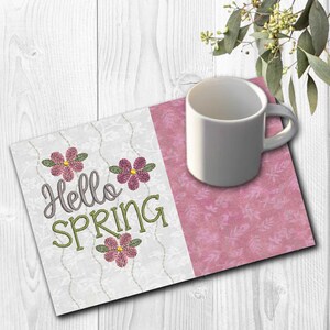 May include: A white ceramic mug rests on a decorative placemat. The placemat features a floral design and the words "Hello SPRING" embroidered with pink and green thread. The background is a white wooden surface.