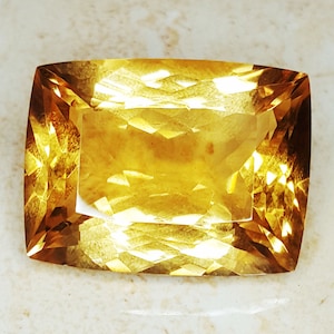 May include: A faceted, rectangular gemstone with rounded corners. The gem is a warm, golden-yellow color, with internal facets creating a sparkling effect. The stone is set against a neutral background, highlighting its color and cut.