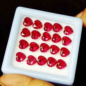 May include: A selection of heart-shaped, faceted, bright red gemstones displayed in a light blue rectangular container. The gemstones are evenly spaced and reflect light. The container is held against a dark background.