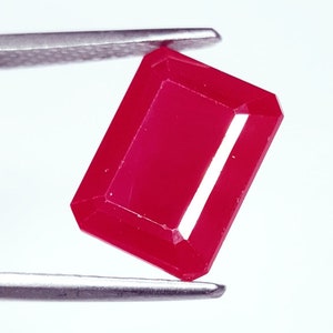 May include: A faceted, rectangular ruby gemstone held by silver tweezers. The ruby is a vibrant, deep red color with sharp edges and a polished surface. The gemstone is set against a bright white background.
