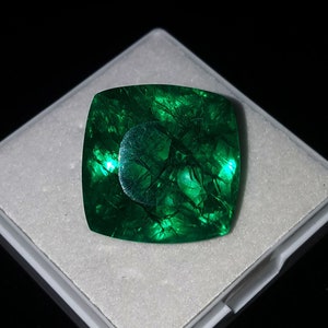 Puede incluir: Una gran esmeralda tallada en forma cuadrada con un tono verde vibrante. La gema está colocada en una caja de exhibición blanca sobre un fondo negro. La superficie de la esmeralda tiene fracturas internas visibles y un área circular reflectante.