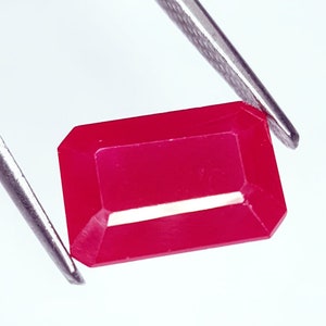 Op de afbeelding: Een gefacetteerde, rechthoekige robijn edelsteen, vastgehouden door een pincet tegen een witte achtergrond. De edelsteen heeft een levendige, dieprode kleur met een gepolijst oppervlak en scherpe randen. De pincet is zilver.