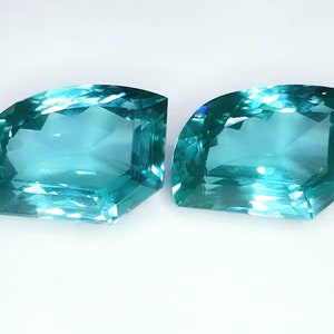 May include: Two teal-coloured, faceted gemstones with a distinctive geometric form. Each gem features a flat, rectangular edge and a curved, semi-circular side. The gems are set against a white backdrop, accentuating their clarity and colour.