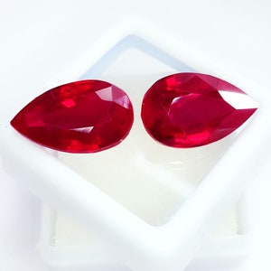 May include: Two faceted, pear-shaped rubies. The gemstones are a deep, vibrant red colour and are set against a white background. The rubies have a high degree of clarity and brilliance, reflecting light. The gems are likely for jewellery making.