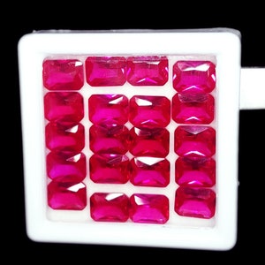 May include: A collection of sixteen rectangular, vibrant pink gemstones displayed in a white tray. The gems are arranged in four rows of four, each with a polished, reflective surface. The background is black.
