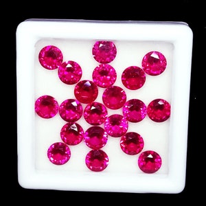 Puede incluir: Un conjunto de quince piedras preciosas redondas, facetadas y de un vibrante color rojo rosado, dispuestas en grupo dentro de una caja de exhibición cuadrada blanca. Cada piedra presenta un corte brillante con múltiples facetas.