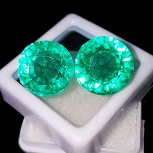 May include: Two round, faceted emerald gemstones with a vibrant green hue are displayed in a white plastic case. The gems have a brilliant cut, reflecting light and showcasing their internal clarity. The background is black, highlighting the gemstones.