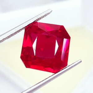 Puede incluir: Un rubí facetado de corte radiante, sujeto con pinzas plateadas. El rubí es de un color rojo intenso y saturado, con bordes definidos y reflejos internos. La gema se muestra sobre un fondo blanco roto.