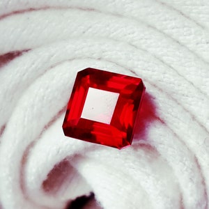 May include: A square-cut, faceted red gemstone with a white square center. The gemstone is set against a white, textured background. The gemstone's edges are sharply defined, reflecting light.