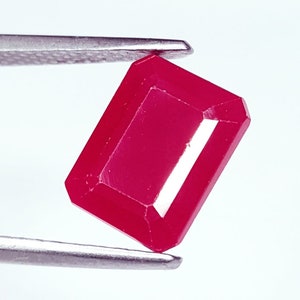 May include: A rectangular-cut ruby gemstone, held by a silver tweezer. The ruby exhibits a deep red colour and is faceted to enhance its brilliance. The gemstone is set against a bright white background.