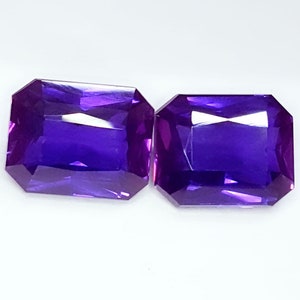 May include: Two rectangular, faceted, deep purple gemstones with a brilliant cut. The gems have a rich, saturated color and are set against a white background. The gems are likely amethyst.