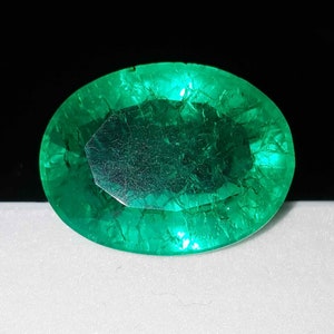 Puede incluir: Una gema de esmeralda de corte ovalado con un tono verde vibrante. La gema muestra fracturas internas y una superficie pulida, sobre un fondo negro. La piedra preciosa es el punto focal, mostrando su belleza natural y claridad.