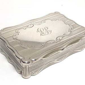 Puede incluir: Una caja rectangular de metal plateado con tapa abatible. La tapa presenta un monograma central grabado con las letras CC rodeado por un borde ornamentado de volutas y rayas. Los bordes de la caja tienen un diseño festoneado.