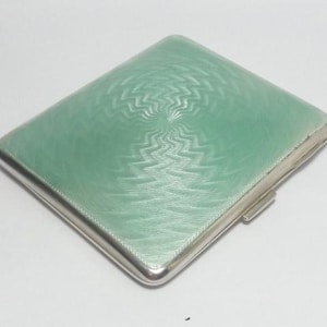 May include: A rectangular, silver-toned cigarette case with a light green, textured surface. The case has a hinged closure and a detailed, swirling pattern on the top. The case is closed and sits on a white surface.